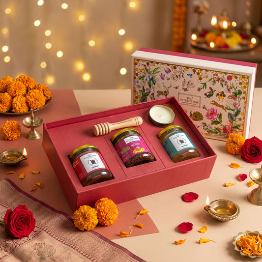 A festive gift set of three Indian honey jars presented in a red box, styled with marigolds, diya lamps, and a floral box on a decorated table.