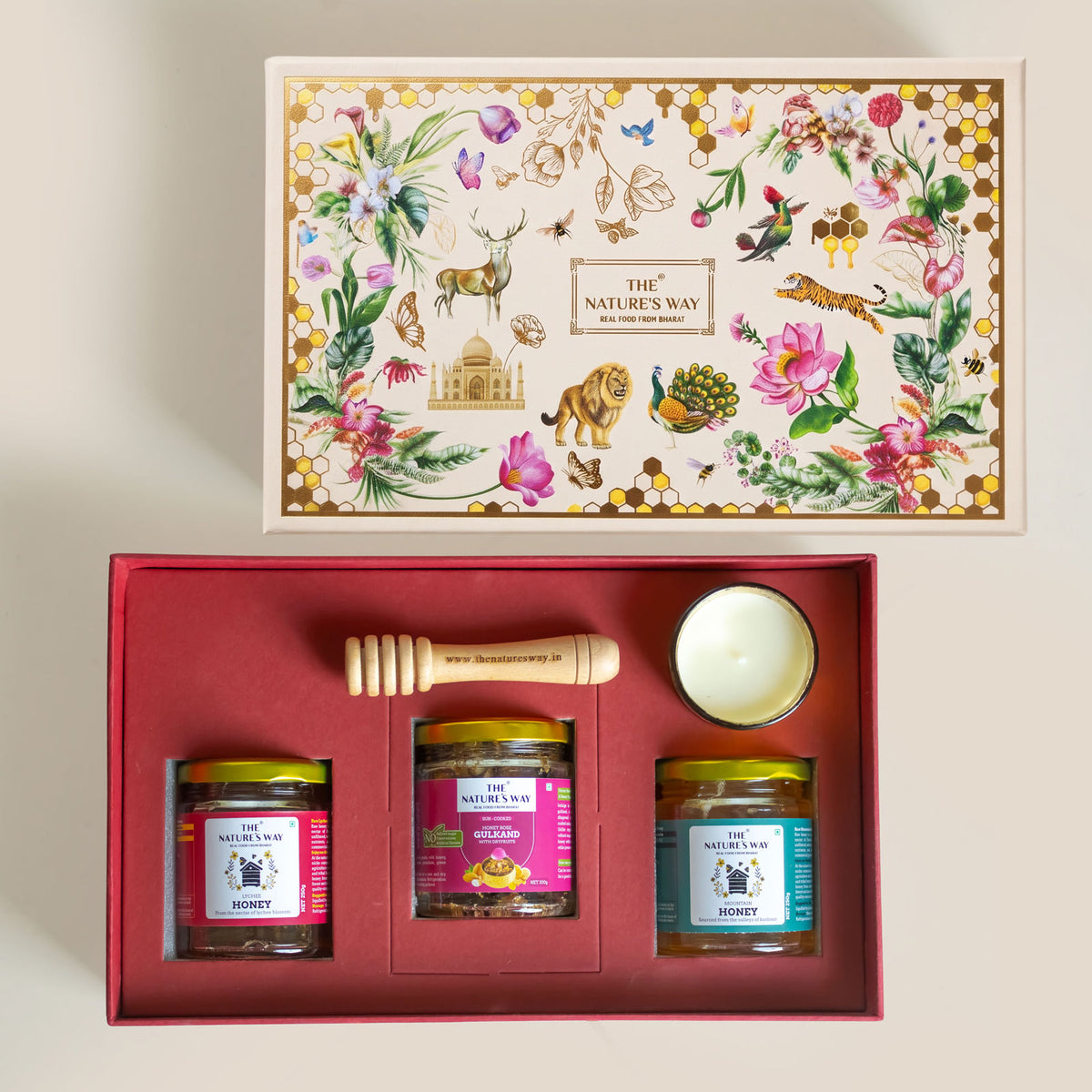 A honey gift set from The Nature's Way, shown open to reveal three glass jars of honey, a wooden honey dipper, and a small white candle in a red presentation box, with the illustrated lid displayed above on a light beige background.