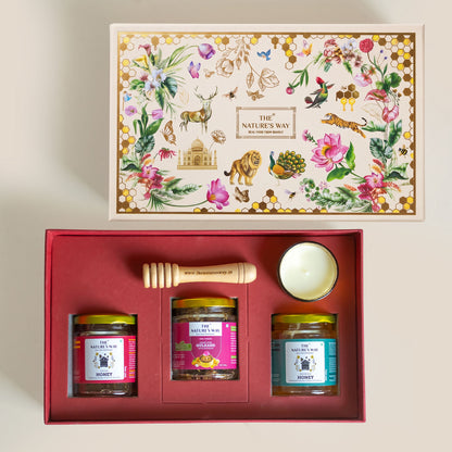 A honey gift set from The Nature's Way, shown open to reveal three glass jars of honey, a wooden honey dipper, and a small white candle in a red presentation box, with the illustrated lid displayed above on a light beige background.