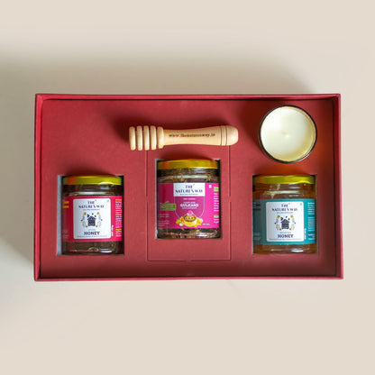A gift set in a red box containing three jars of honey, a wooden honey dipper, and a small white candle, photographed from above on a light beige background.